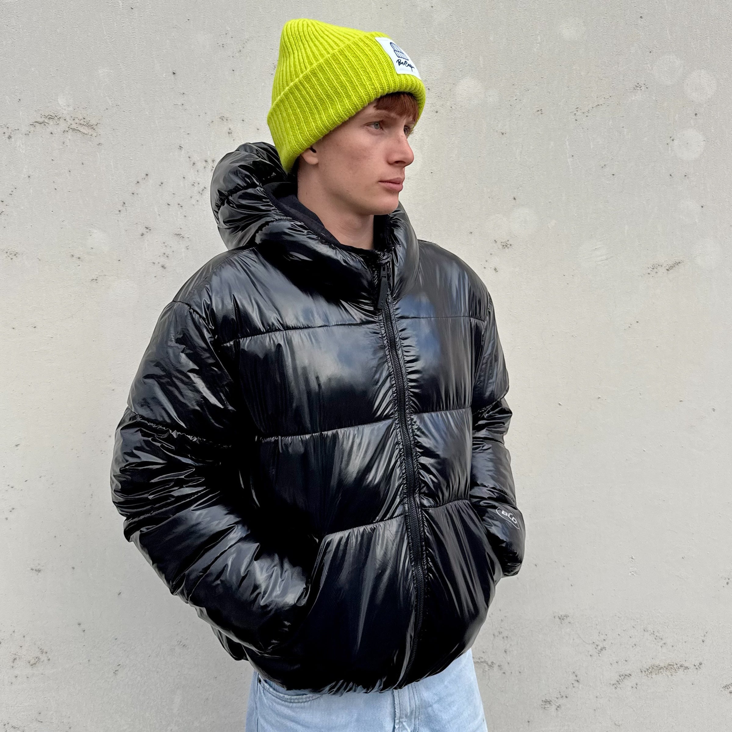 Giovane uomo con giacca black puffer e cuffia neon gialla, stile streetwear, sfondo grigio.